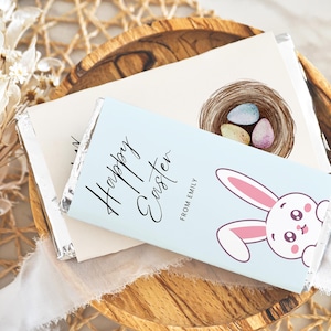 Può includere: Un involucro di cioccolato blu e bianco con il testo "Buona Pasqua" e una simpatica illustrazione di un coniglietto. L'involucro include anche il testo "Da Emily".