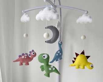 May include: A white baby mobile with a gray crescent moon, white clouds, and four felt dinosaur toys in green, blue, yellow, and pink.