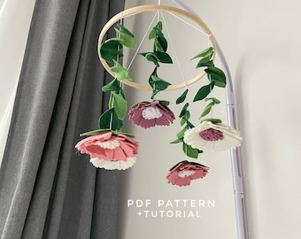 Peut inclure: Un mobile en bois avec des fleurs et des feuilles en feutre. Les fleurs sont roses, blanches et vertes. Le mobile est suspendu à un support blanc. Le texte "PDF PATTERN + TUTORIAL" est imprimé sous le mobile.