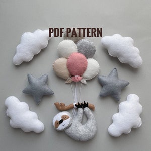 Puede incluir: Patrón PDF para un móvil de fieltro con un perezoso colgado, nubes, estrellas y globos. El perezoso es gris, las nubes son blancas, las estrellas son grises y los globos son rosas, blancos y beige.