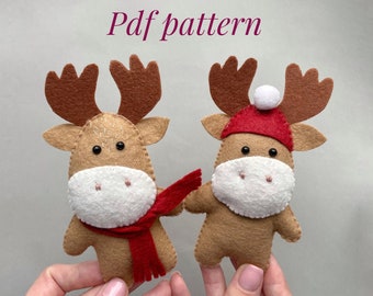 May include: Two felt reindeer ornaments, one wearing a red hat and the other with a red scarf. The reindeer are brown with white faces and have stitched details. The text "Pdf pattern" is written above the reindeer.