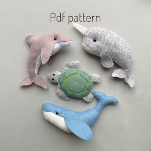 May include: A set of four felt animal toys: a pink dolphin, a gray narwhal, a green sea turtle, and a blue whale. The toys are arranged on a gray background. Pdf pattern.