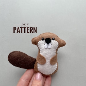 May include: A brown felt beaver with a white belly and black eyes. The beaver is holding a brown felt tail. The text "PDF PATTERN" is above the beaver.