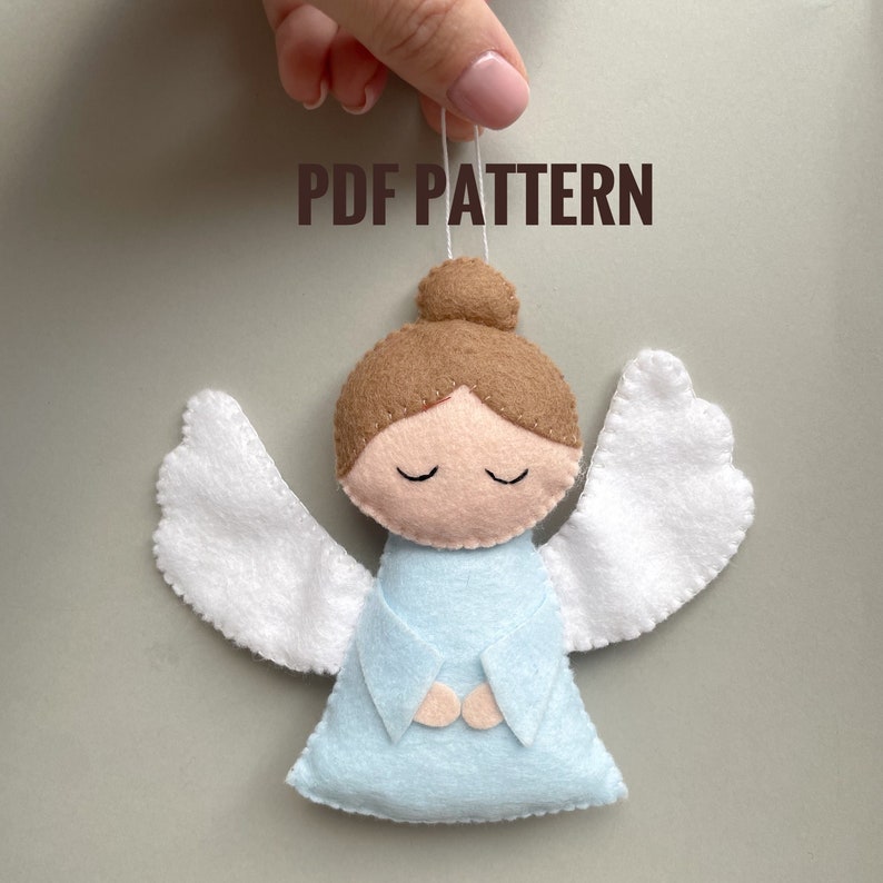 Puede incluir: Patr&oacute;n PDF para un adorno de &aacute;ngel de fieltro. El &aacute;ngel es azul claro con alas blancas y un mo&ntilde;o de pelo marr&oacute;n. El &aacute;ngel tiene los ojos cerrados y las manos juntas en oraci&oacute;n.