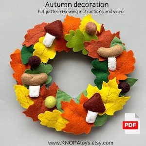 Puede incluir: Una corona de otoño de fieltro con setas marrones, bellotas y hojas en tonos naranja, amarillo y verde. La corona está decorada con tallos de fieltro blanco y un icono PDF. El texto "Autumn decoration" y "Pdf pattern+sewing instructions and video" están en la parte superior de la imagen. El texto "www.KNOPAToys.etsy.com" está en la parte inferior de la imagen.