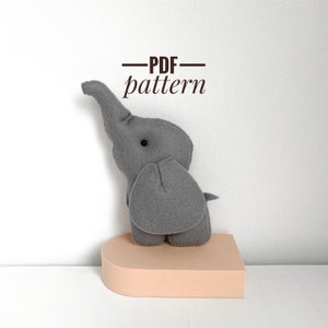 Può includere: Un giocattolo di peluche di elefante in feltro grigio con occhi neri, in piedi su una base rosa. Il testo "PDF pattern" è scritto sopra il giocattolo.