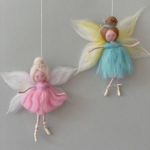 May include: Two felt fairy ornaments hanging from white strings. The fairy on the left has pink wings, a pink dress, and blonde hair. The fairy on the right has yellow wings, a blue dress, and brown hair.