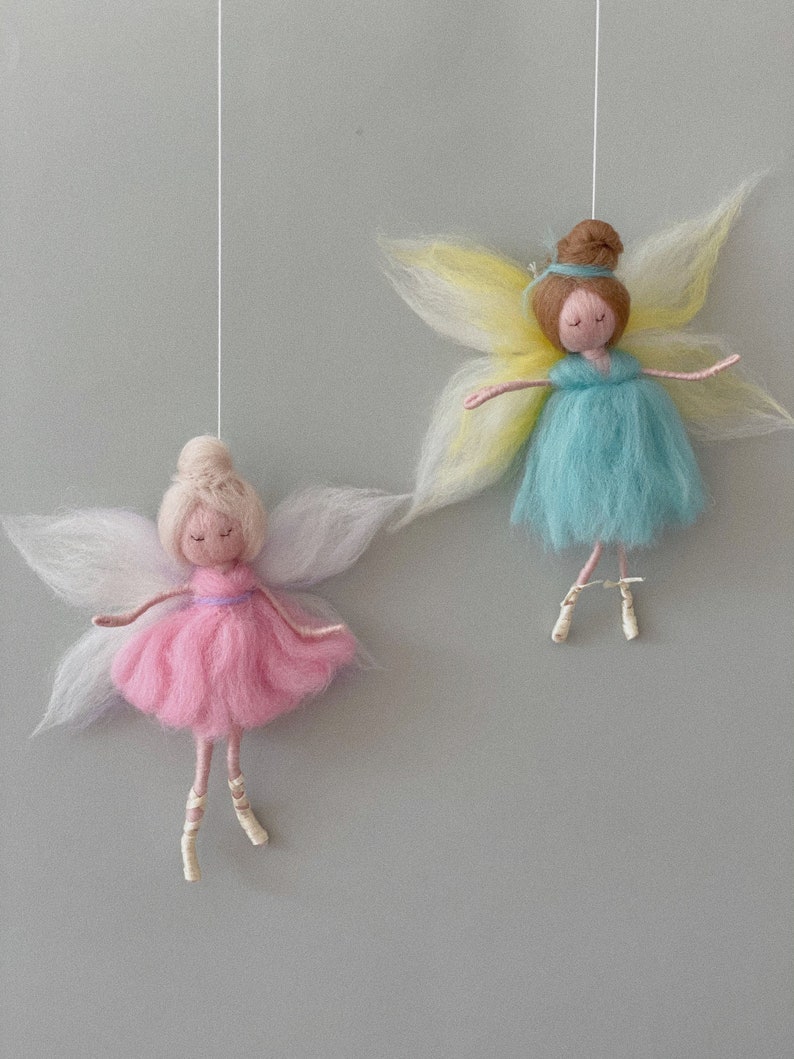 May include: Two felt fairy ornaments hanging from white strings. The fairy on the left has pink wings, a pink dress, and blonde hair. The fairy on the right has yellow wings, a blue dress, and brown hair.