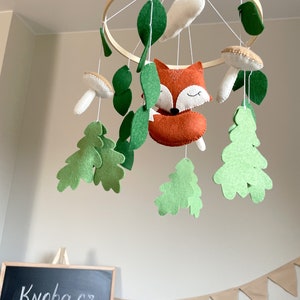 May include: A wooden hoop mobile with felt woodland animals and leaves. The mobile features a fox, mushrooms, and oak leaves in shades of green, brown, and white.