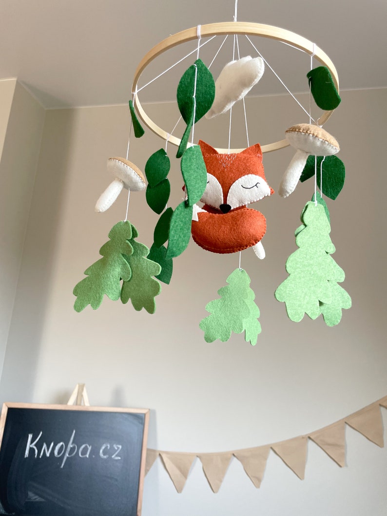 May include: A wooden hoop mobile with felt woodland animals and leaves. The mobile features a fox, mushrooms, and oak leaves in shades of green, brown, and white.