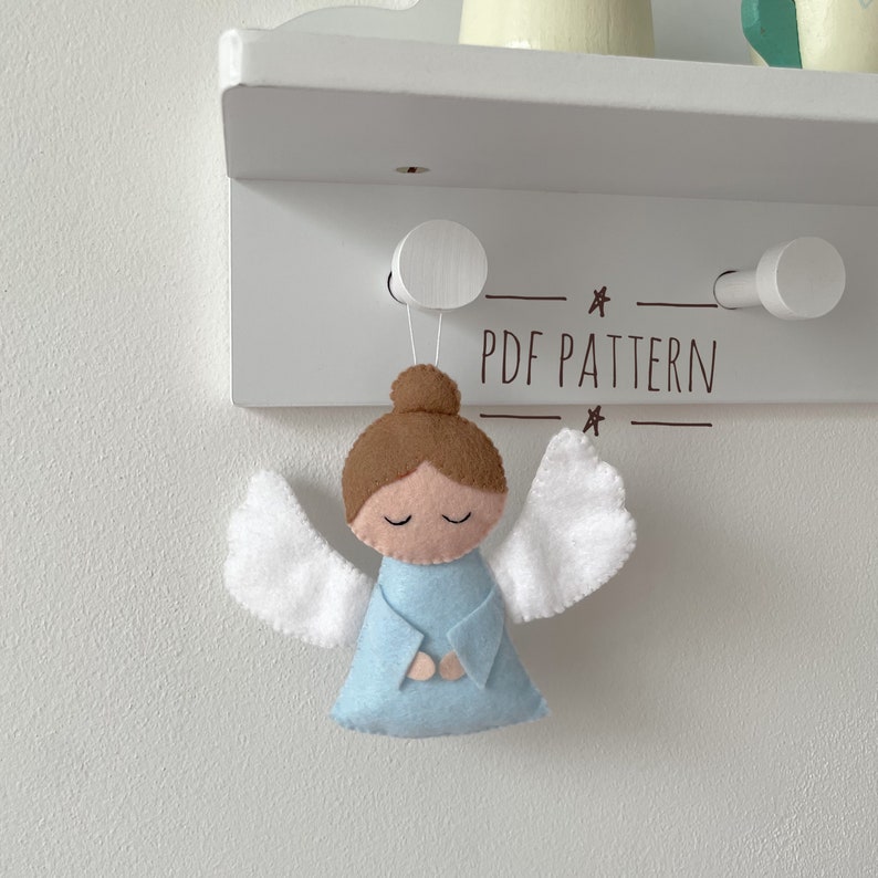 Puede incluir: Un adorno de &aacute;ngel de fieltro con cabello casta&ntilde;o, vestido azul y alas blancas. El &aacute;ngel est&aacute; colgado de un estante blanco con el texto "PDF PATTERN" impreso en &eacute;l.