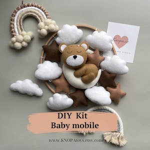 Può includere: Kit fai-da-te per un mobile per bambini con un orso marrone che dorme su una mezzaluna bianca, circondato da nuvole bianche e stelle marroni. Il kit include un cerchio di legno, pezzi di feltro e istruzioni.