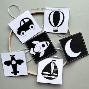 May include: Black and white felt cards with images of a car, hot air balloon, rocket, moon, airplane, and sailboat. The cards are hanging from a wooden hoop.