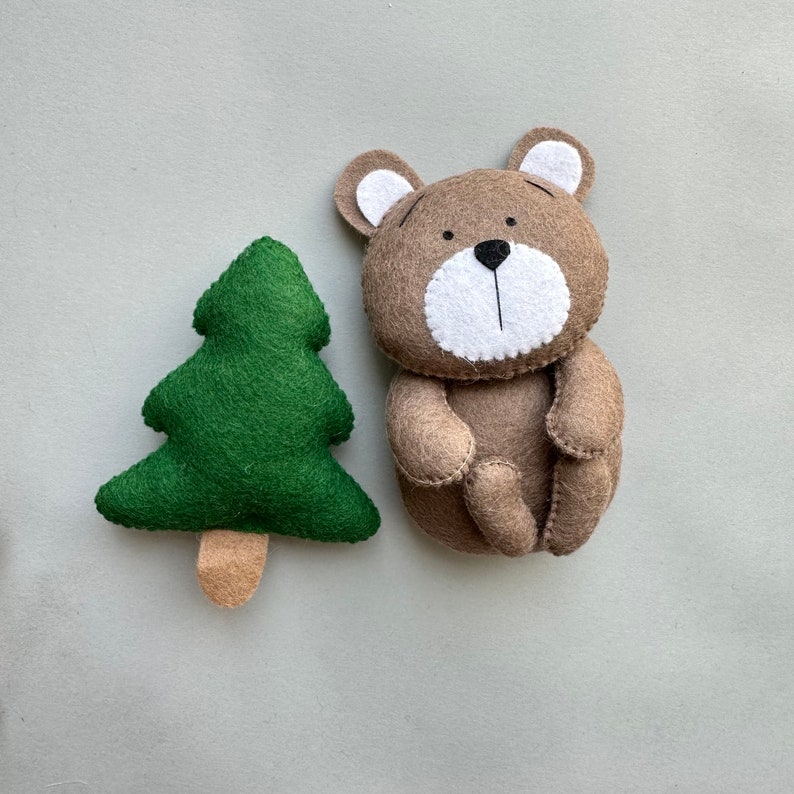 May include: A brown felt bear with white felt ears, nose, and mouth. The bear is sitting with its legs crossed. Next to the bear is a green felt Christmas tree with a brown felt trunk.