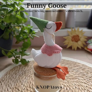 Könnte beinhalten: Eine handgefertigte Gans aus Filz mit grünem Hut und rosa Kragen, die auf einem Glas mit Korken sitzt. Die Gans hat orangefarbene Füße und einen weißen Körper. Der Text "Funny Goose" und "KNOPAToys" sind sichtbar.
