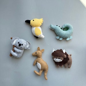 May include: Five felt animal toys: a yellow and white cockatoo, a gray koala holding a branch, a brown kangaroo, a brown and white bison, and a light blue alligator.