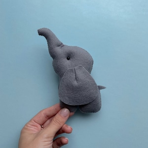K&ouml;nnte beinhalten: Ein graues Filz-Elefantenspielzeug mit einem schwarzen Knopfauge. Der Elefant steht auf einer hellblauen Oberfl&auml;che.