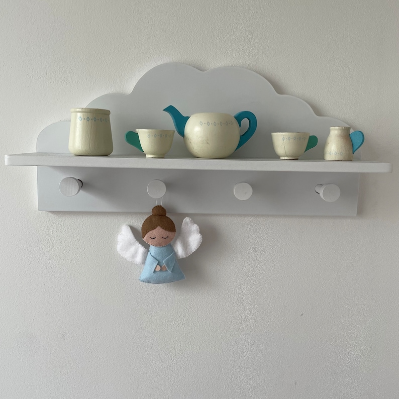 Puede incluir: Estante blanco en forma de nube con ganchos, que incluye una tetera azul, dos tazas de t&eacute; blancas, una jarra de crema blanca y un azucarero blanco. Un adorno de &aacute;ngel de fieltro azul cuelga de uno de los ganchos.