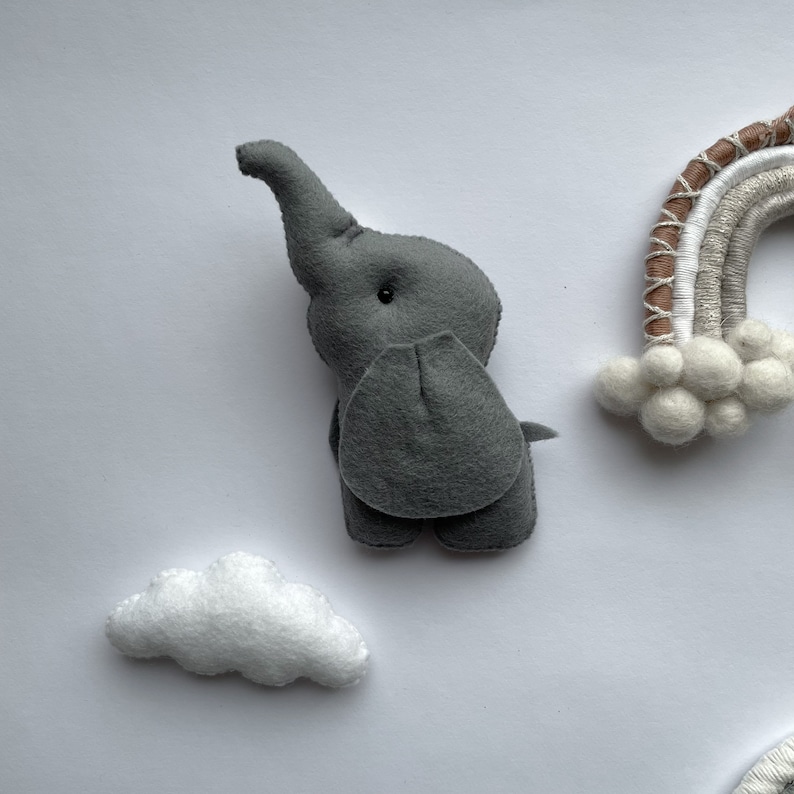 K&ouml;nnte beinhalten: Ein graues Filz-Elefantenspielzeug mit einem langen R&uuml;ssel und einer wei&szlig;en Filzwolke. Der Elefant steht auf einer wei&szlig;en Oberfl&auml;che.