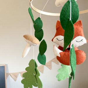 May include: A wooden hoop mobile with felt woodland animal and leaf decorations. The mobile features a fox, a mushroom, and leaves in shades of green and brown.