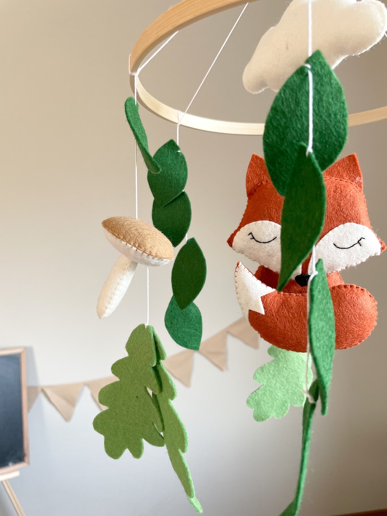 May include: A wooden hoop mobile with felt woodland animal and leaf decorations. The mobile features a fox, a mushroom, and leaves in shades of green and brown.