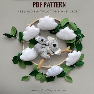 Può includere: Un modello PDF per un mobile in feltro con un koala, nuvole e foglie. Il mobile è appeso a un cerchio di legno. Il modello include istruzioni di cucito e un video.