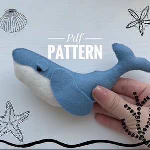 May include: A blue felt whale plush toy with a white belly and a fin. The whale is being held in a hand against a white background. The text "Pdf Pattern" is written in black on the white background.