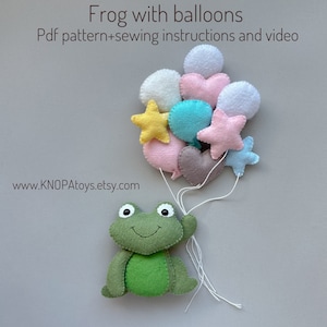 Puede incluir: Una rana de fieltro verde sosteniendo un montón de globos de fieltro de colores. Los globos tienen forma de corazones, estrellas y círculos. Los globos están unidos a la rana con un hilo blanco. El fondo es de color gris claro. El texto "www.KNOPAtoys.etsy.com" está en la parte inferior de la imagen.