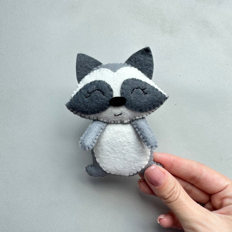 May include: A gray and white felt raccoon toy with a smiling face. The toy has black eyes and a black nose.