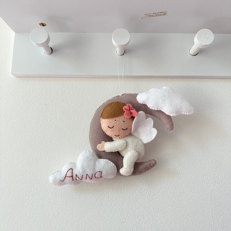 Pu&ograve; includere: Un angelo di feltro con capelli castani e un fiore rosa che dorme su una luna crescente grigia con nuvole bianche. Il nome "Anna" &egrave; ricamato su una delle nuvole.