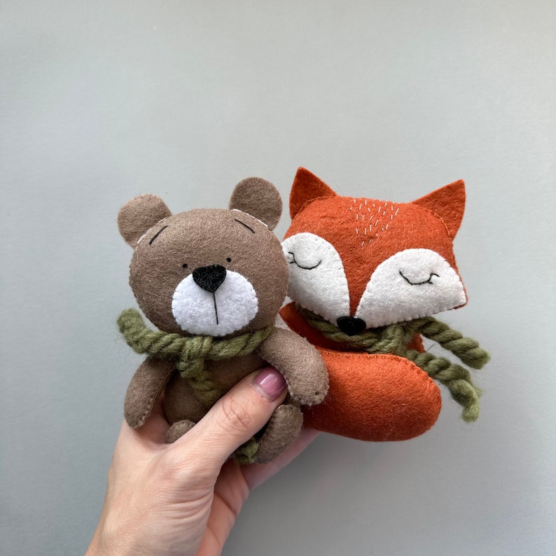 May include: A brown bear and an orange fox plush toy with green scarves. The toys are made of felt and have embroidered details.