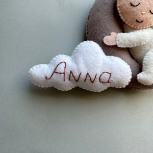 Pu&ograve; includere: Una nuvola di feltro bianco con il nome "Anna" ricamato in filo marrone. La nuvola &egrave; attaccata a una luna crescente di feltro grigio con una piccola bambola di feltro che dorme su di essa.