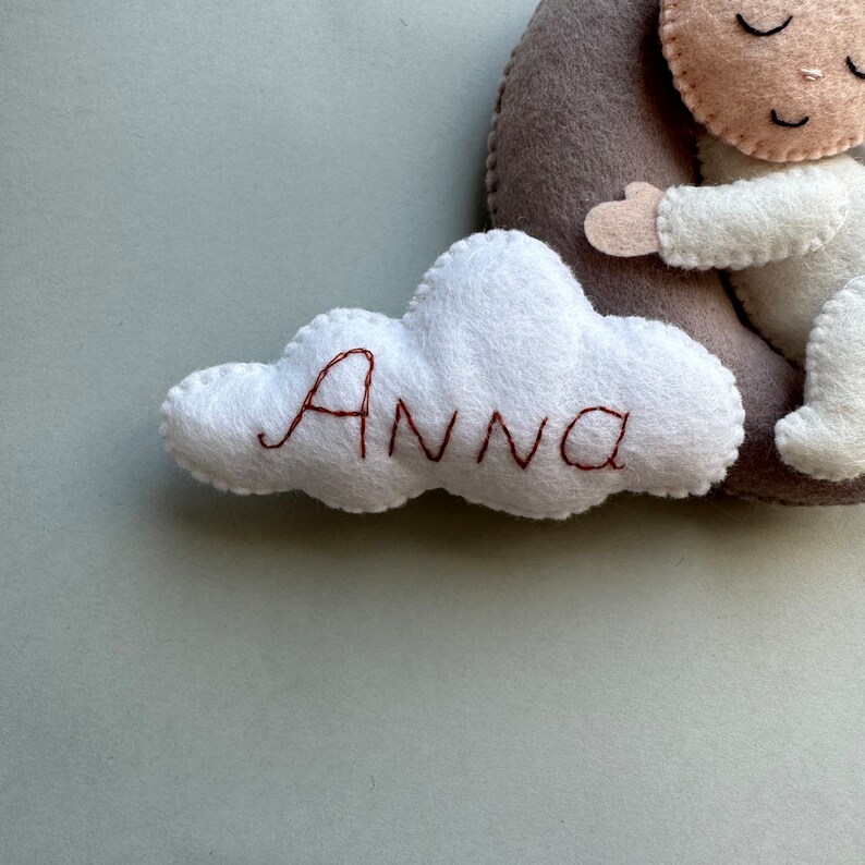 Pu&ograve; includere: Una nuvola di feltro bianco con il nome "Anna" ricamato in filo marrone. La nuvola &egrave; attaccata a una luna crescente di feltro grigio con una piccola bambola di feltro che dorme su di essa.