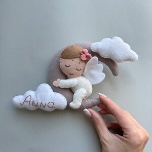 Pu&ograve; includere: Un angelo di feltro con i capelli castani e un fiore rosa in testa sta dormendo su una luna crescente grigia. L'angelo indossa un vestito bianco e ha ali bianche. La luna &egrave; circondata da due nuvole bianche, una delle quali ha il nome "Anna" ricamato.