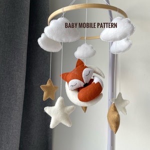 May include: A white, brown, and orange felt baby mobile with a fox sleeping on a crescent moon, three white stars, and two brown stars. The mobile is hanging from a wooden ring and says "BABY MOBILE PATTERN" on the ring.
