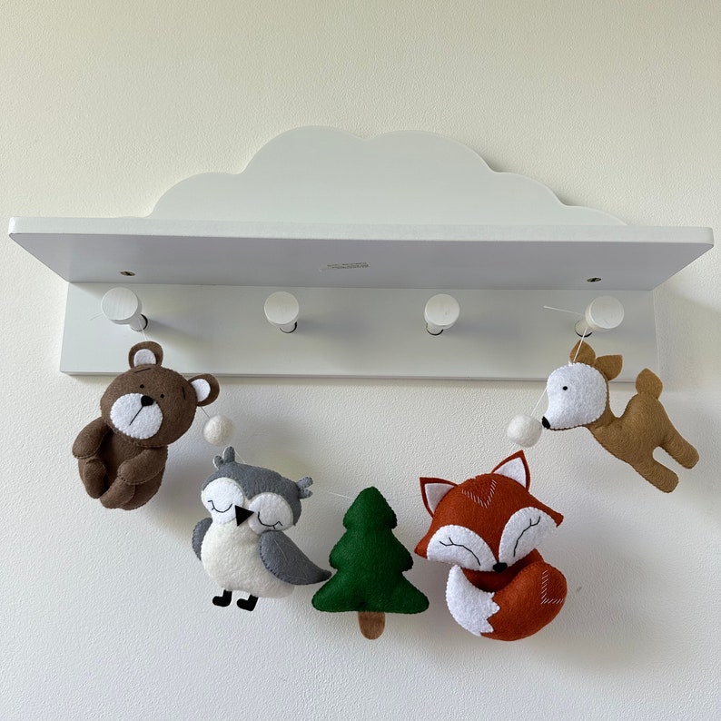 May include: A white shelf with five hooks, each holding a felt animal hanging from a string. The animals are a brown bear, a gray owl, a green tree, a red fox, and a brown deer.