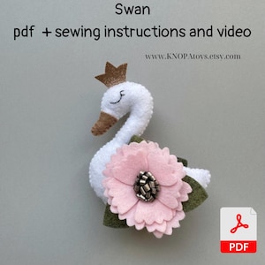 Pode incluir: Um cisne de feltro branco com uma coroa dourada, uma flor de feltro rosa e folhas de feltro verde. O cisne está segurando a flor em seu bico. A imagem inclui o texto "Swan" e "pdf + sewing instructions and video".