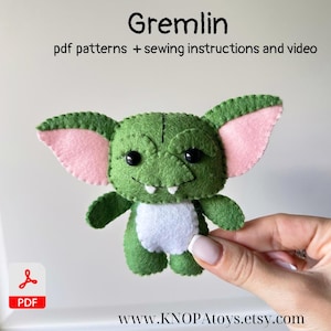 May include: A handmade green felt Gremlin doll with pink inner ears, black eyes, and white teeth. The doll has a white belly and small arms. The image includes the text "Gremlin" and "pdf patterns + sewing instructions and video".