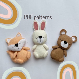 May include: Three felt animal toys: a brown bear, a white bunny with pink ears, and an orange fox. The toys have embroidered eyes and mouths. The text "PDF patterns" is written above the toys.