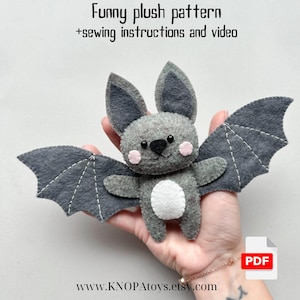 Può includere: Un giocattolo di peluche a forma di pipistrello in feltro grigio con pancia bianca e guance rosa. Il pipistrello ha grandi ali ed è tenuto in una mano. L'immagine include il testo "Funny plush pattern +sewing instructions and video" e "PDF".