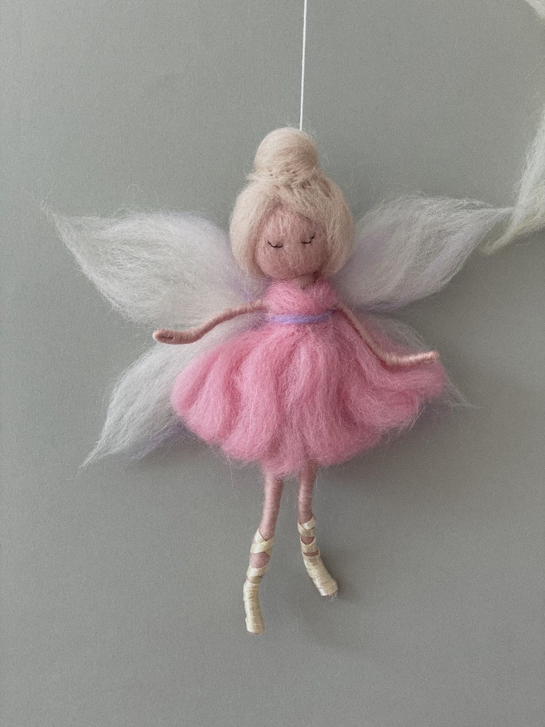 May include: A pink felt fairy doll with white and purple wings, blonde hair, and a purple ribbon around her waist. She is wearing white ballet shoes and has her eyes closed.