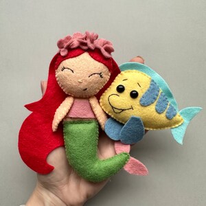 May include: A felt mermaid doll with red hair, a pink top, and a green tail. The mermaid is holding a yellow felt fish with blue fins.