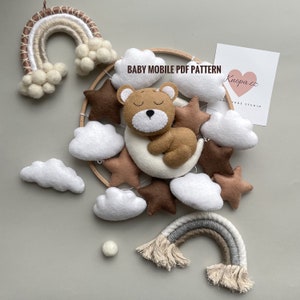 May include: A brown felt bear sleeping on a white crescent moon, surrounded by brown felt stars and white felt clouds. The mobile is hanging from a wooden hoop and includes a rainbow made of yarn.