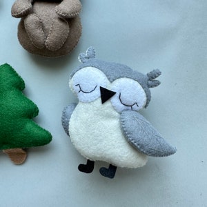 May include: A gray and white felt owl with black eyes and a black beak. The owl is sitting on a light blue surface.