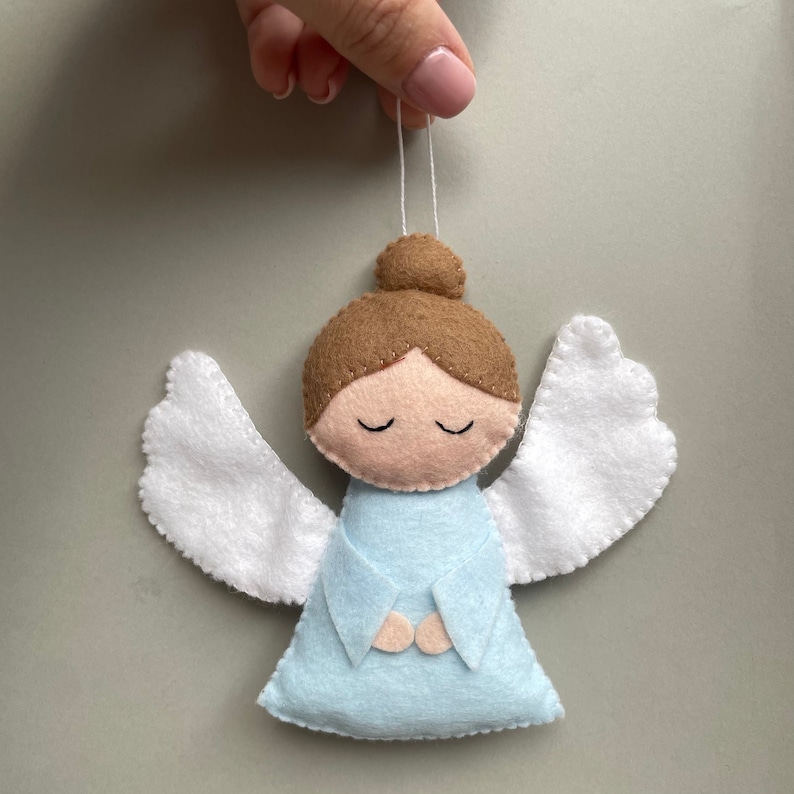 Puede incluir: Un adorno de &aacute;ngel de fieltro con alas blancas, cabello casta&ntilde;o y un vestido azul claro. El &aacute;ngel tiene los ojos cerrados y las manos juntas en oraci&oacute;n.