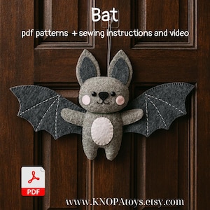 May include: A gray felt bat with dark gray wings and a white belly, hanging on a wooden door. The bat has pink cheeks and a black nose and eyes. Text above the bat reads "Bat" and "pdf patterns + sewing instructions and video".