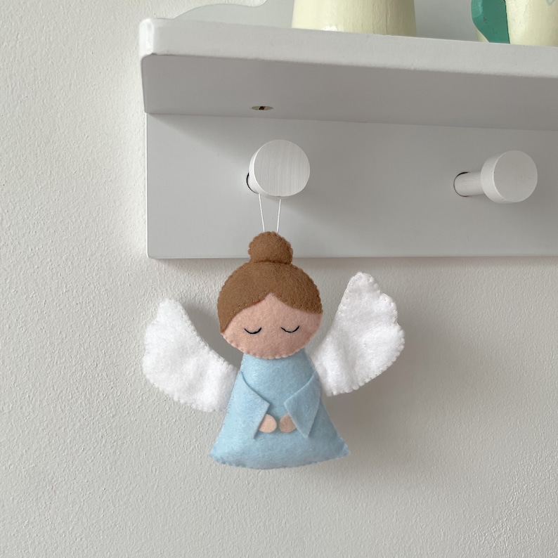 Puede incluir: Un adorno de &aacute;ngel de fieltro con cabello casta&ntilde;o, un vestido azul claro y alas blancas. El &aacute;ngel est&aacute; colgado de un gancho blanco en un estante blanco.