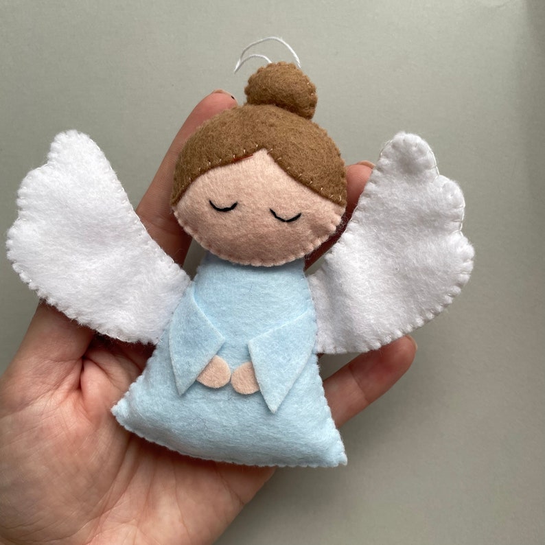 Puede incluir: Un adorno de &aacute;ngel de fieltro con alas blancas, cabello casta&ntilde;o y un vestido azul claro. El &aacute;ngel tiene los ojos cerrados y las manos juntas en oraci&oacute;n.