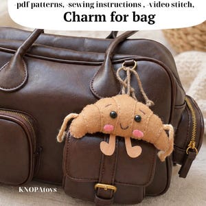 May include: A brown leather bag with a small pocket and a gold buckle. A felt croissant charm with a smiling face and small legs hangs from the bag. The text "Charm for bag" is above the charm.