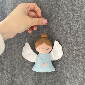 Puede incluir: Un adorno de &aacute;ngel de fieltro con alas blancas, cabello casta&ntilde;o y un vestido azul claro. El &aacute;ngel tiene los ojos cerrados y las manos juntas en oraci&oacute;n.
